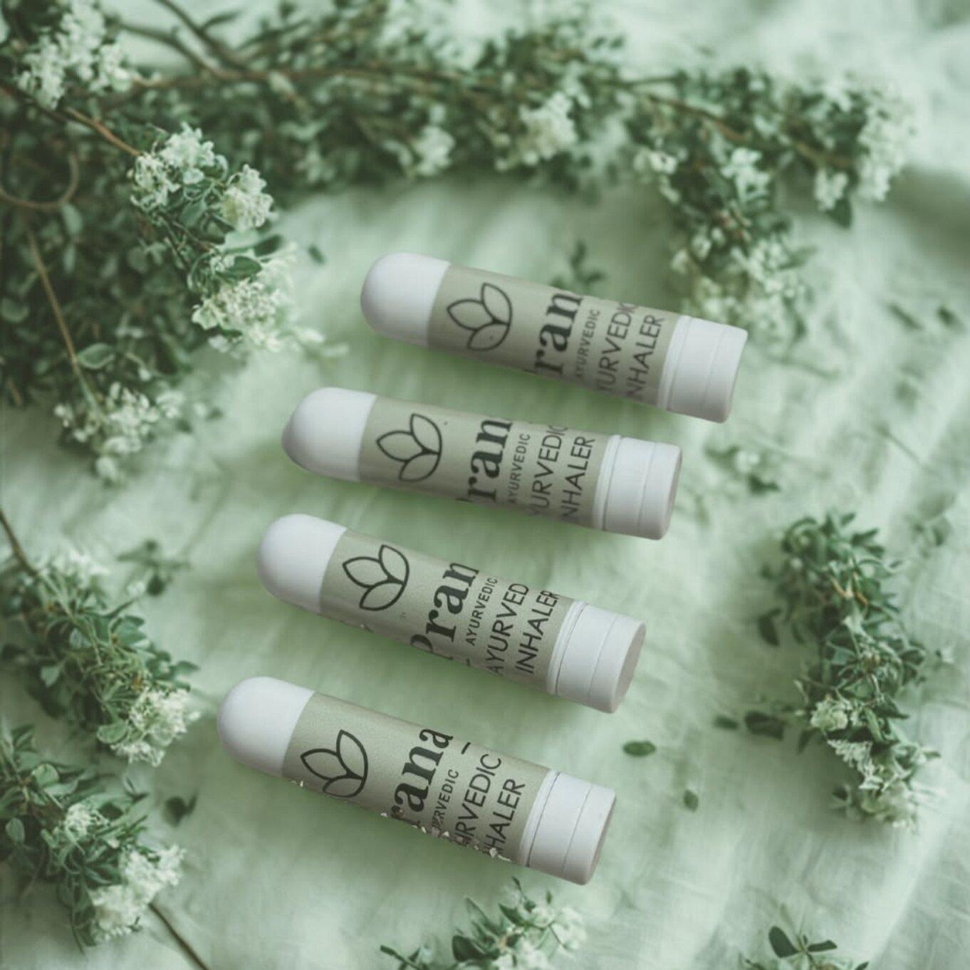 Cuatro inhaladores Prana Ayurvedic sobre una tela verde, entre ramas con flores blancas